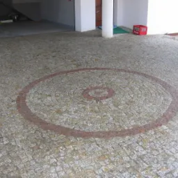 Podjazd wyłożony drobną kostką granitową w odcieniach szarości, z dekoracyjnym wzorem koła w kolorze czerwono-brązowym, otoczony kolumnami i zadaszeniem.